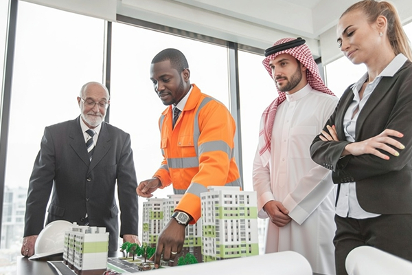Four business professionals, including a man in an orange safety jacket, stand around a table with architectural models of buildings, discussing plans for facilitating foreign investors in Kenya in a modern office with large windows in the background.