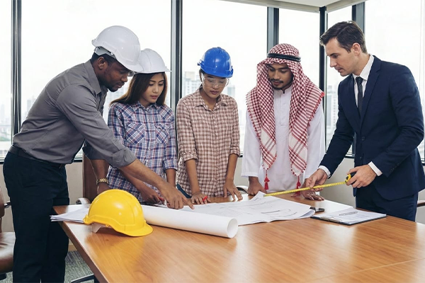 Five people, including engineers wearing hard hats and diverse attire, stand around a table in an office, examining blueprints and using a tape measure—showcasing the best in construction project management in Kenya. Large windows overlook a cityscape.